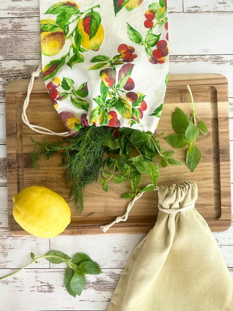 May include: A wooden cutting board with fresh herbs, a lemon, and two drawstring bags. One bag is filled with dill and mint, and the other is empty. The bags are made of linen and cotton. The bag with herbs has a white fabric with a colorful fruit pattern.