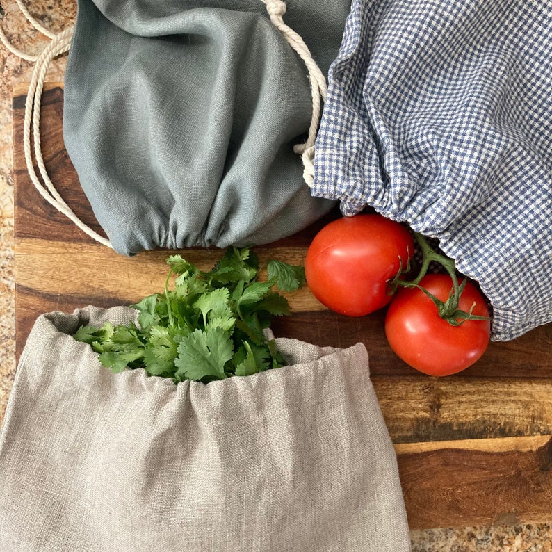 May include: Three reusable produce bags in different colors and patterns sit on a wooden cutting board. One is gray, one is beige, and the third is blue and white checkered. Fresh tomatoes and cilantro are visible.