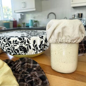 Może przedstawiać: Zestaw materiałowych przykrywek do misek, obejmujący beżową osłonę na szklanym słoiku, osłonę w czarno-biały wz&oacute;r kwiatowy na ż&oacute;łtej misce oraz brązową osłonę w kratę, wszystkie wykończone elastycznymi brzegami dla lepszego dopasowania.
