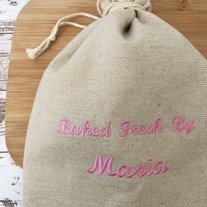 May include: A beige linen drawstring bag with the embroidered text "Baked Fresh By Maria" in pink script. The bag is closed with a natural-colored rope and sits on a wooden cutting board.