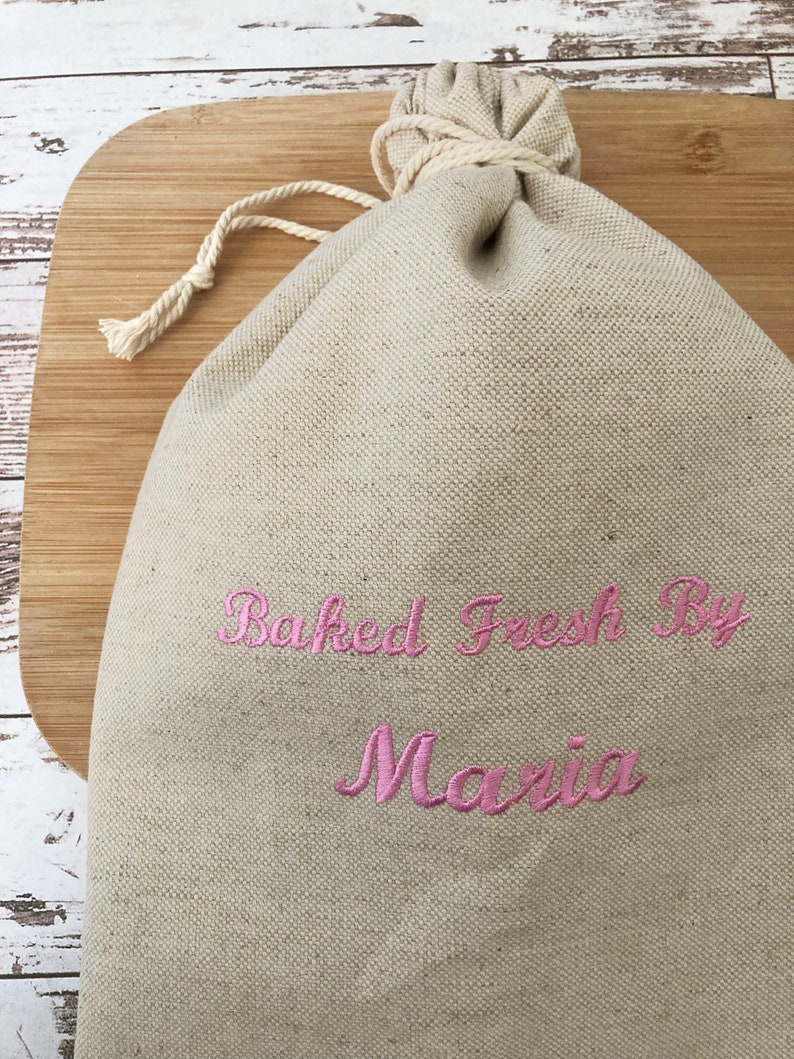 May include: A beige linen drawstring bag with the embroidered text "Baked Fresh By Maria" in pink script. The bag is closed with a natural-colored rope and sits on a wooden cutting board.