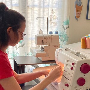May include: A white Janome sewing machine and a white Singer serger machine are positioned on a table. The text Handmade in Los Angeles, CA, USA is visible at the top. The sewing machine features pink dials and a printed stitch guide on the front.