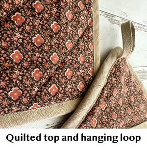 May include: Two quilted oven mitts with a floral pattern in shades of brown, red, and pink. The mitts have a quilted top and a hanging loop made of a natural-coloured fabric. The text "Quilted top and hanging loop" is visible.