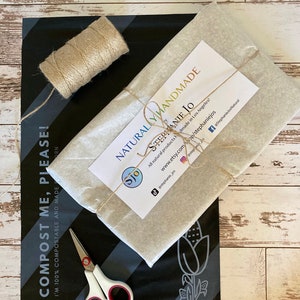 Może przedstawiać: Prostokątna paczka owinięta w jasnobeżowy papier z białą etykietą z napisem NATURALLY HANDMADE STEPHANIE JO, przewiązana sznurkiem. Obok znajduje się czarna kompostowalna koperta wysyłkowa, szpulka sznurka oraz białe nożyczki z czerwonymi uchwytami.