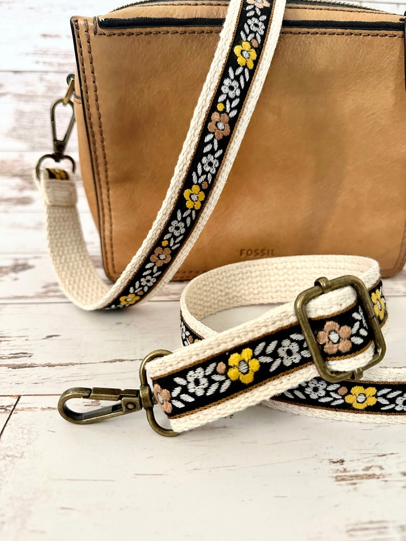 May include: A tan leather Fossil crossbody bag with a detachable, adjustable woven strap. The strap features a black background with a repeating pattern of yellow and white flowers, green leaves, and tan accents, finished with antique brass hardware.