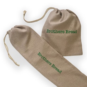 May include: Two beige linen bread bags with drawstring closures. The bags are different sizes, one long and narrow, the other shorter and wider. The words "Brothers Bread" are embroidered in green on both bags. The bags are made of a natural-colored fabric.