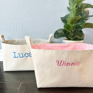 Op de afbeelding: Twee canvas opbergmanden met gepersonaliseerde namen. De mand links is blauw en zegt "Luca" en de mand rechts is roze en zegt "Winnie".