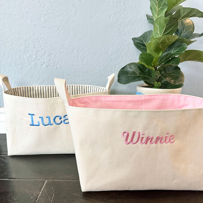Op de afbeelding: Twee canvas opbergmanden met gepersonaliseerde namen. De mand links is blauw en zegt "Luca" en de mand rechts is roze en zegt "Winnie".