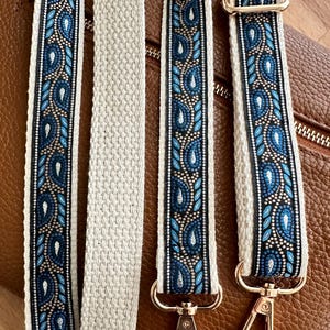 May include: A replacement bag strap featuring a woven pattern of blue and silver paisley shapes on a black background, bordered by cream-colored fabric. The strap includes gold-toned metal swivel lobster clasps and an adjustable slider buckle.