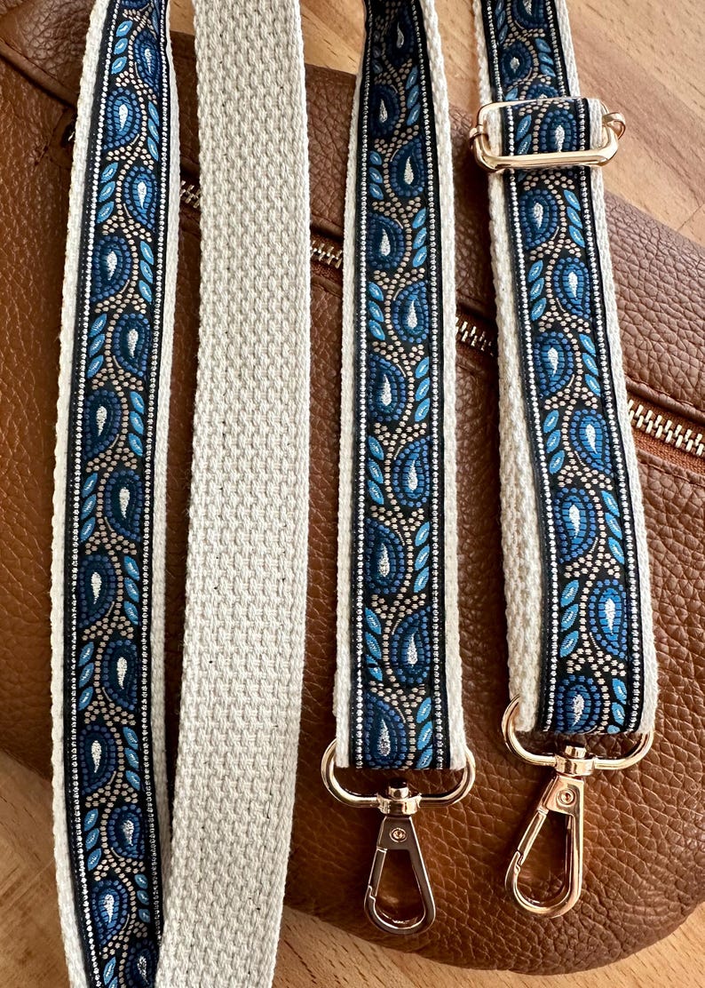 May include: A replacement bag strap featuring a woven pattern of blue and silver paisley shapes on a black background, bordered by cream-colored fabric. The strap includes gold-toned metal swivel lobster clasps and an adjustable slider buckle.