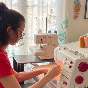 Op de afbeelding: Een persoon naait op een witte Janome naaimachine met een roze spoelhouder. De machine staat op een houten tafel met een Singer overlockmachine op de achtergrond. De persoon draagt een rood shirt en gebruikt beide handen om de stof te geleiden.
