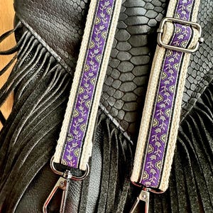 May include: A black bag with a textured snakeskin pattern and long fringe details, featuring two adjustable straps. The straps are made of cream-colored fabric with a central purple band decorated with a repeating floral pattern and silver hardware.