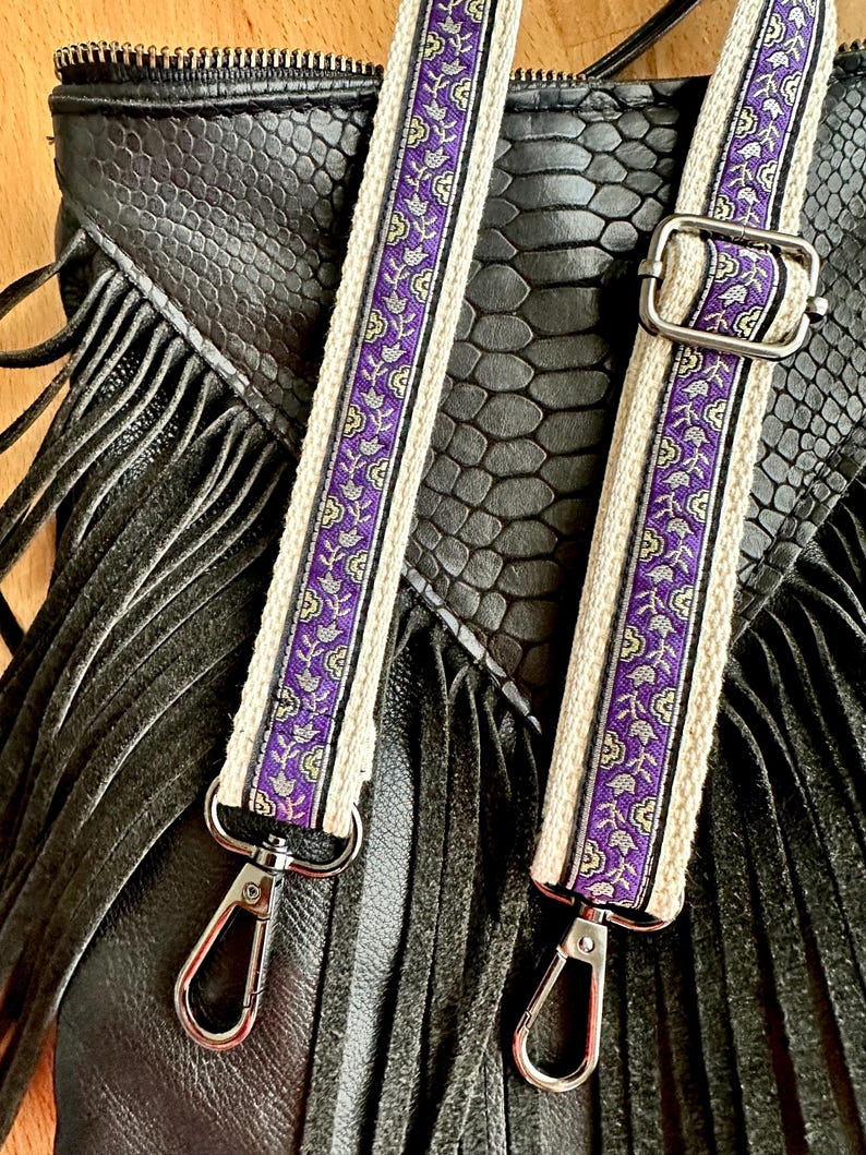May include: A black bag with a textured snakeskin pattern and long fringe details, featuring two adjustable straps. The straps are made of cream-colored fabric with a central purple band decorated with a repeating floral pattern and silver hardware.