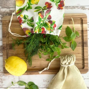 Puede incluir: Una tabla de cortar de madera con hierbas frescas, un lim&oacute;n y dos bolsas con cord&oacute;n. Una bolsa est&aacute; llena de eneldo y menta, y la otra est&aacute; vac&iacute;a. Las bolsas son de lino y algod&oacute;n. La bolsa con hierbas tiene una tela blanca con un estampado de frutas coloridas.