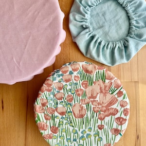 May include: Three round fabric bowl covers with elasticated edges. One cover is solid light pink, one is solid light blue, and one features a floral pattern with pink flowers, green stems, and small blue accents on a white background.