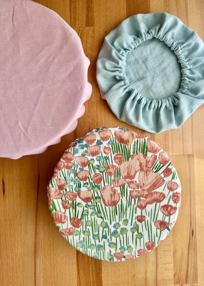 May include: Three round fabric bowl covers with elasticated edges. One cover is solid light pink, one is solid light blue, and one features a floral pattern with pink flowers, green stems, and small blue accents on a white background.