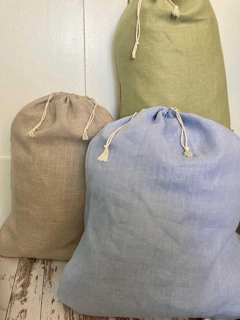 May include: Three drawstring bags made of a textured fabric in light blue, beige, and sage green. Each bag features a gathered top closure secured with a white twisted cord, creating a soft, rounded shape for storage and organization purposes.