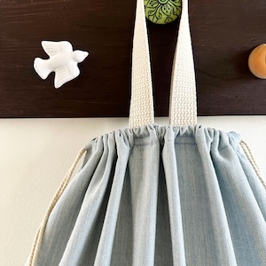 May include: A light blue drawstring bag with cream-colored straps hangs from a wooden hook. The bag is made of a textured fabric. A white ceramic bird and a green floral knob are also visible.