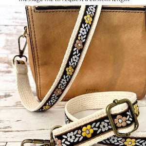 May include: A 1 inch wide bag strap with an adjustable length up to 54 inches. The strap features a black center band with embroidered yellow and light brown flowers and white leaves, bordered by cream-colored fabric and antique brass hardware.