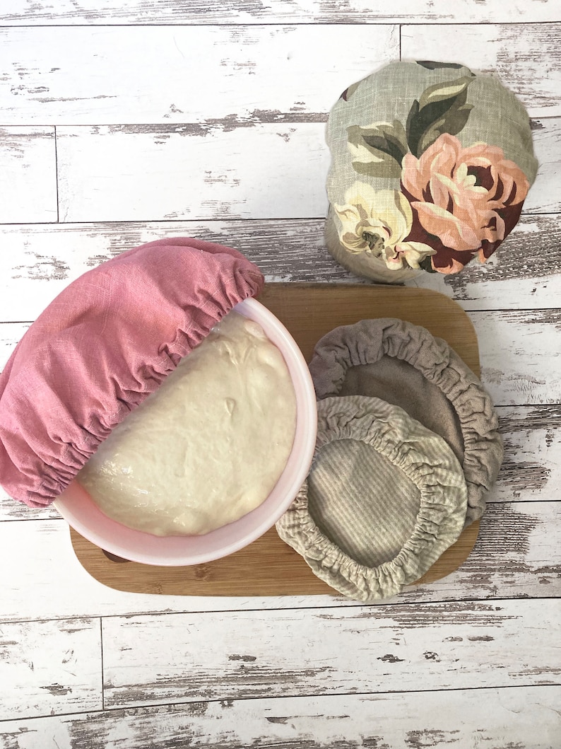 May include: A set of four fabric bowl covers with elasticated edges. One cover is solid dusty pink and placed over a bowl of dough. Another features a floral print with roses. Two additional covers are in neutral beige and a subtle striped pattern.