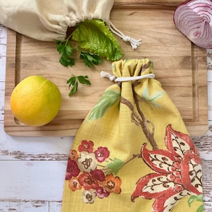 May include: A yellow drawstring bag featuring a floral pattern in shades of red, orange, and green, paired with a plain cream-colored drawstring bag. Both bags are made of fabric and include white rope closures for securing the contents inside.