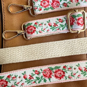 May include: Three bag straps featuring a white background with a repeating red rose and green leaf floral pattern. Two straps include gold-toned metal lobster clasps and an adjustable slider, while the third is a plain, textured beige woven strap.