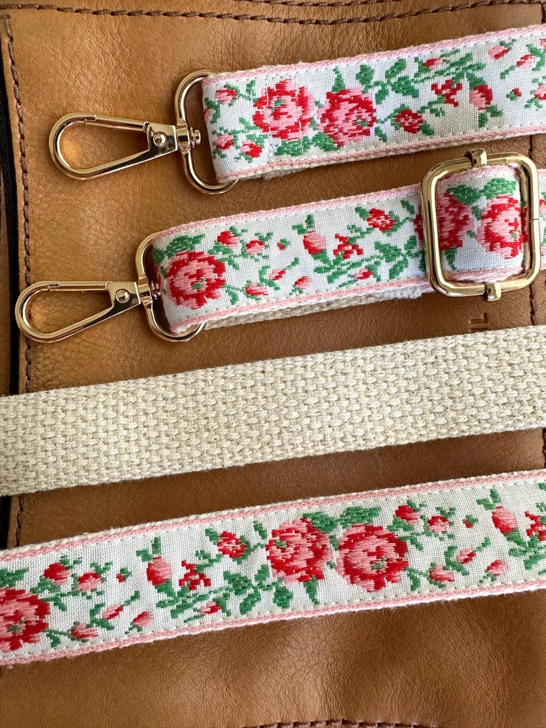 May include: Three bag straps featuring a white background with a repeating red rose and green leaf floral pattern. Two straps include gold-toned metal lobster clasps and an adjustable slider, while the third is a plain, textured beige woven strap.