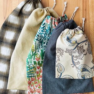May include: A set of five fabric drawstring bags in various patterns and colors. The collection includes a brown and white plaid bag, a solid tan bag, a white bag with a green and red floral print, a solid dark gray bag, and a cream bag with a blue paisley print.