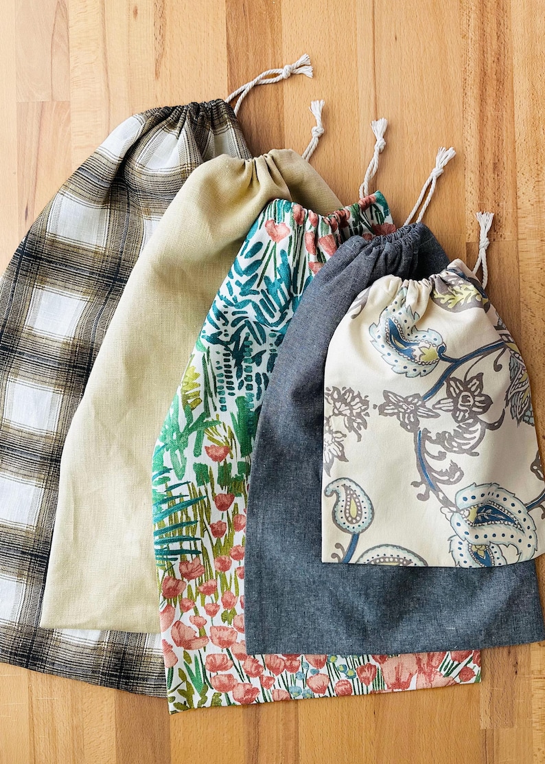 May include: A set of five fabric drawstring bags in various patterns and colors. The collection includes a brown and white plaid bag, a solid tan bag, a white bag with a green and red floral print, a solid dark gray bag, and a cream bag with a blue paisley print.