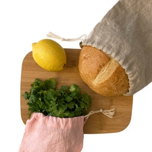 May include: A wooden cutting board holds a loaf of bread in a beige drawstring bag, a yellow lemon, and a bunch of cilantro in a pink drawstring bag. The bread is round and golden brown. The cilantro is green and leafy.