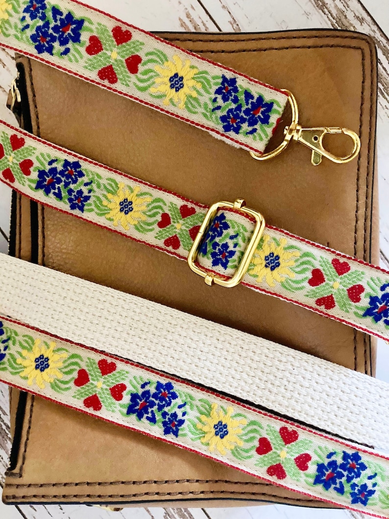 May include: A woven bag strap featuring a cream-colored base with a repeating floral pattern in blue, yellow, green, and red. The strap includes gold-toned metal hardware, including a lobster clasp and an adjustable slider buckle for length.