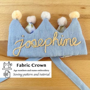 May include: A light blue fabric crown with the name "Josephine" embroidered in a gold thread. The crown has five peaks, each topped with a small, fluffy, cream-colored pom-pom. The text "Fabric Crown" is visible.