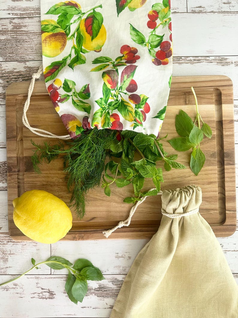 May include: A wooden cutting board with fresh herbs, a lemon, and two drawstring bags. One bag is filled with dill and mint, and the other is empty. The bags are made of linen and cotton. The filled bag has a white fabric with a fruit and floral pattern.