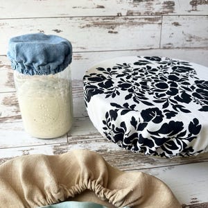 May include: A collection of fabric bowl covers including a blue cover on a glass jar, a white cover with a black floral pattern, and two additional covers in solid tan and light blue, all featuring elasticated edges for a secure fit.