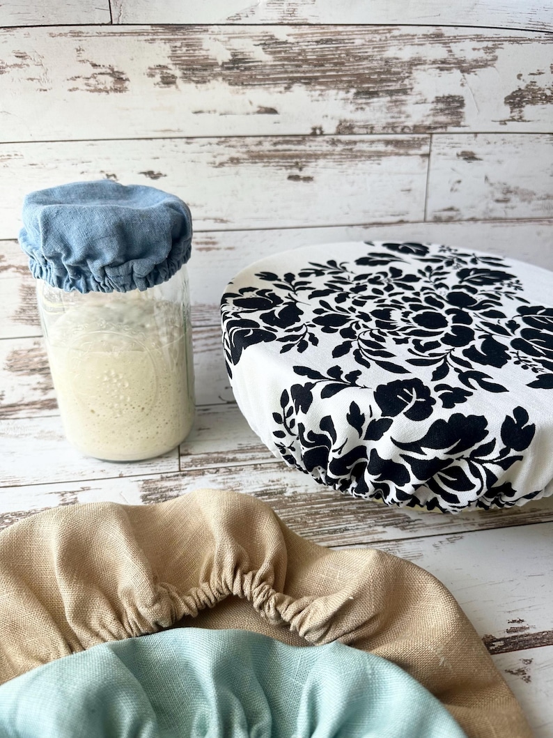 May include: A collection of fabric bowl covers including a blue cover on a glass jar, a white cover with a black floral pattern, and two additional covers in solid tan and light blue, all featuring elasticated edges for a secure fit.
