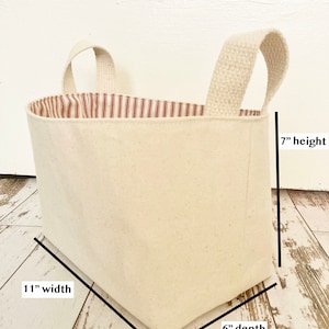 Op de afbeelding: Een witte canvas opbergmand met een rode en witte gestreepte stoffen voering. De mand heeft twee stoffen handvatten en meet 28 cm breed, 15 cm diep en 18 cm hoog.