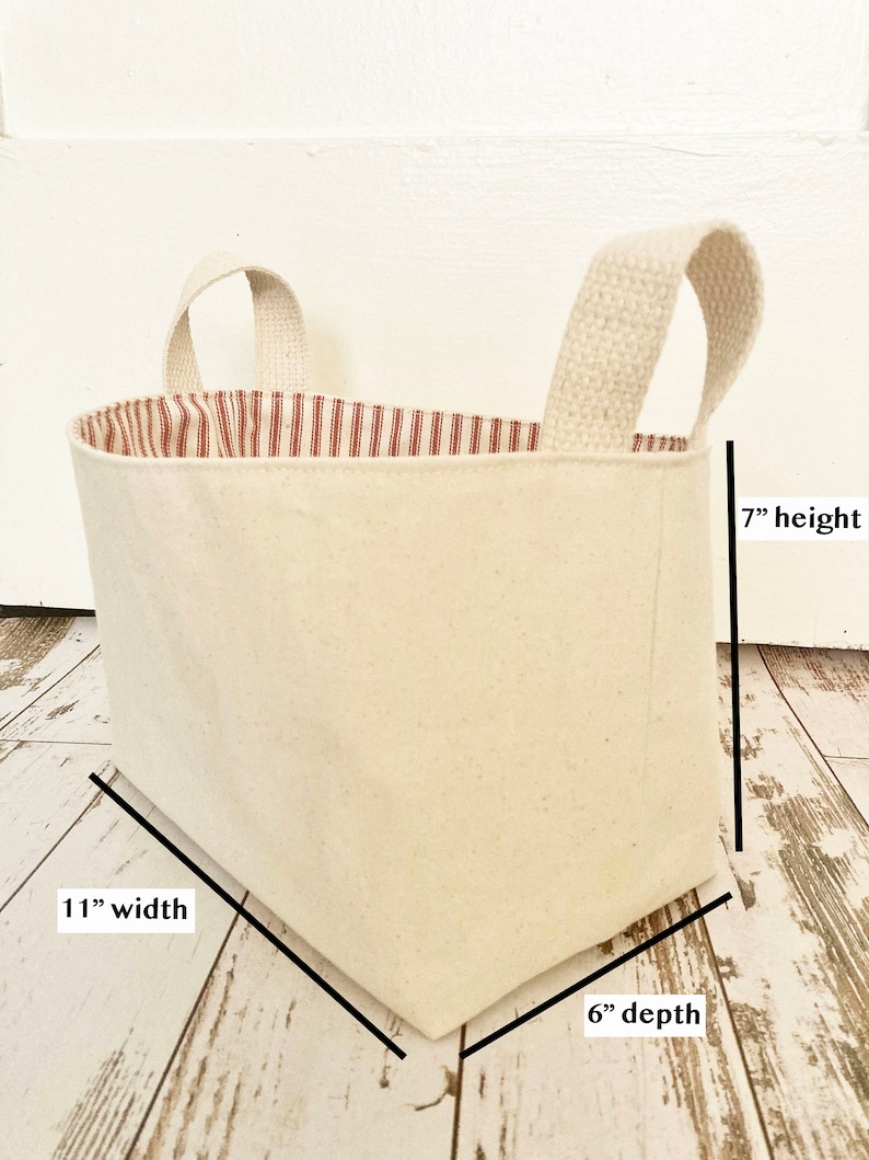 Op de afbeelding: Een witte canvas opbergmand met een rode en witte gestreepte stoffen voering. De mand heeft twee stoffen handvatten en meet 28 cm breed, 15 cm diep en 18 cm hoog.