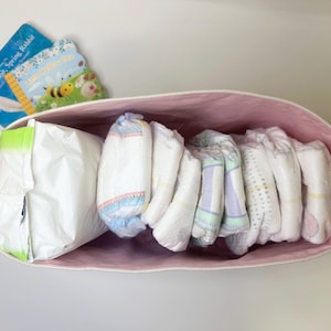 Op de afbeelding: Een roze canvas opbergmand met een witte touwgreep, gevuld met verschillende witte babyluiers en een witte plastic zak. De mand staat op een wit oppervlak.