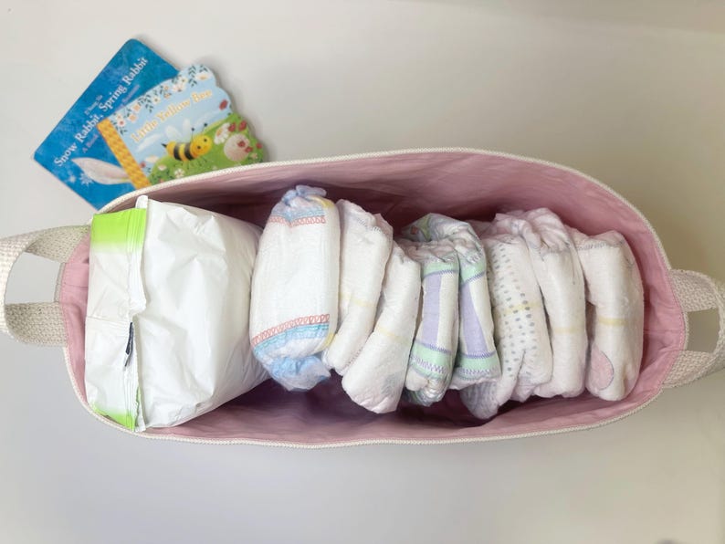 Op de afbeelding: Een roze canvas opbergmand met een witte touwgreep, gevuld met verschillende witte babyluiers en een witte plastic zak. De mand staat op een wit oppervlak.