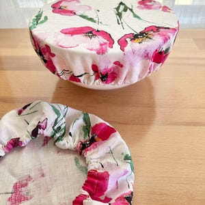 May include: Two round fabric bowl covers featuring a white background with a watercolour-style floral print in shades of bright pink, magenta, and green. One cover is fitted over a bowl, while the other lies flat, both showing elasticated edges.