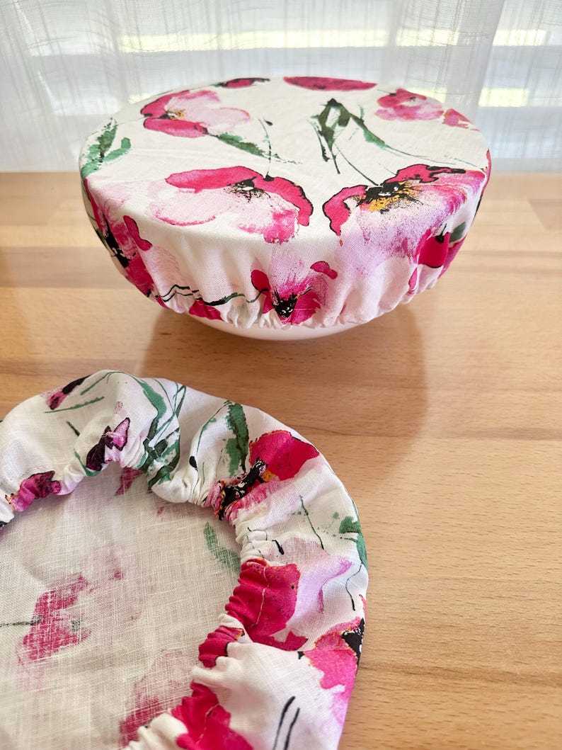 May include: Two round fabric bowl covers featuring a white background with a watercolour-style floral print in shades of bright pink, magenta, and green. One cover is fitted over a bowl, while the other lies flat, both showing elasticated edges.