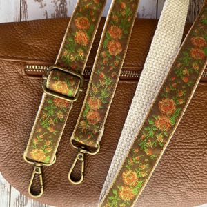 May include: A brown leather bag featuring two detachable bag straps. The straps are made of a brown fabric with a woven floral pattern in orange and green, featuring antique brass-colored metal hardware including lobster clasps and sliders.