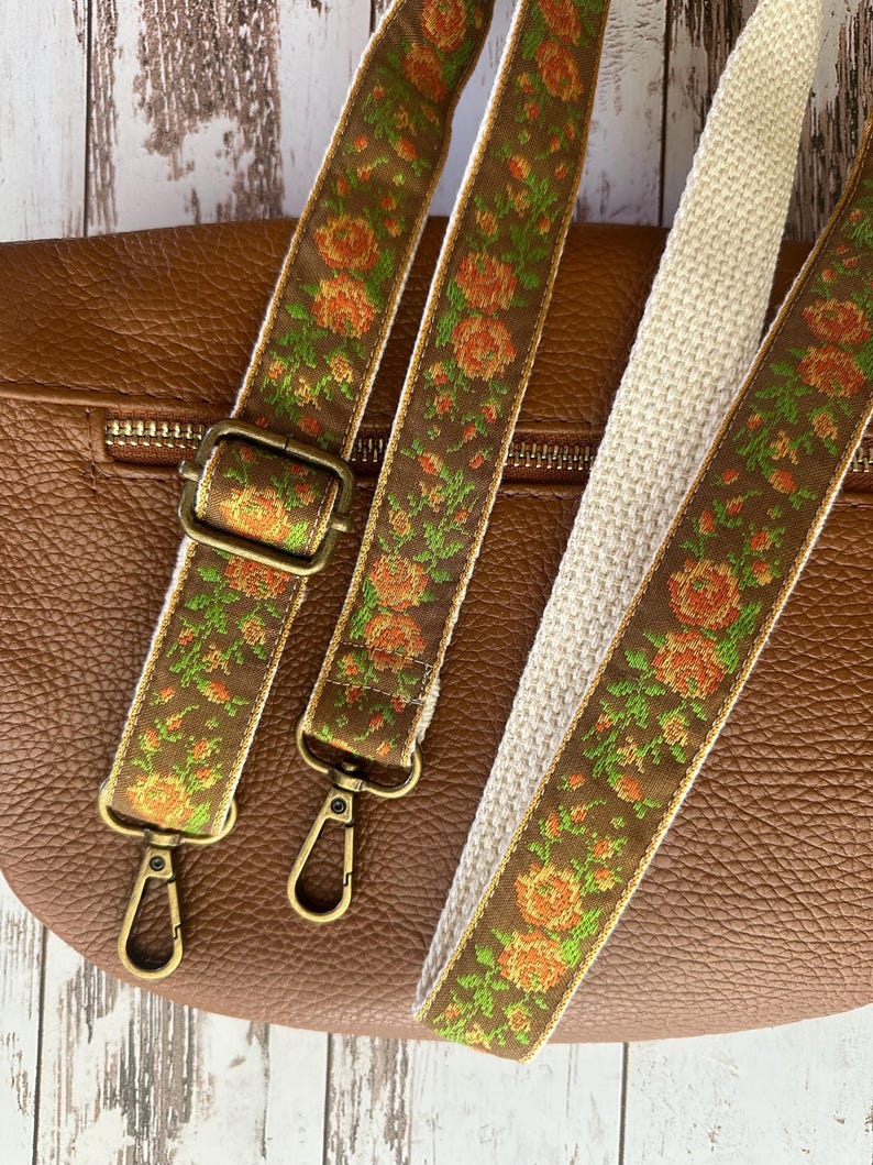 May include: A brown leather bag featuring two detachable bag straps. The straps are made of a brown fabric with a woven floral pattern in orange and green, featuring antique brass-colored metal hardware including lobster clasps and sliders.