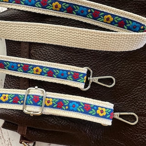 May include: A dark brown leather bag featuring a detachable, adjustable shoulder strap. The strap is made of cream-colored woven fabric with a central blue ribbon embroidered with a repeating pattern of red, yellow, and blue flowers and green leaves.