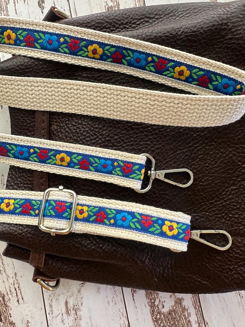 May include: A dark brown leather bag featuring a detachable, adjustable shoulder strap. The strap is made of cream-colored woven fabric with a central blue ribbon embroidered with a repeating pattern of red, yellow, and blue flowers and green leaves.