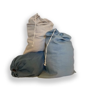 May include: Three fabric drawstring bags in varying shades of blue and light gray. Each bag features a simple woven cord closure at the top, allowing the opening to be cinched shut. The bags have a soft, textured appearance and are arranged together.