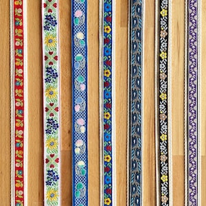 May include: Seven vertical strips of decorative woven ribbon featuring distinct patterns: red floral, red hearts, blue cherries with a grid, blue floral, blue paisley, black floral, and purple floral. Each ribbon has a cream-coloured border.