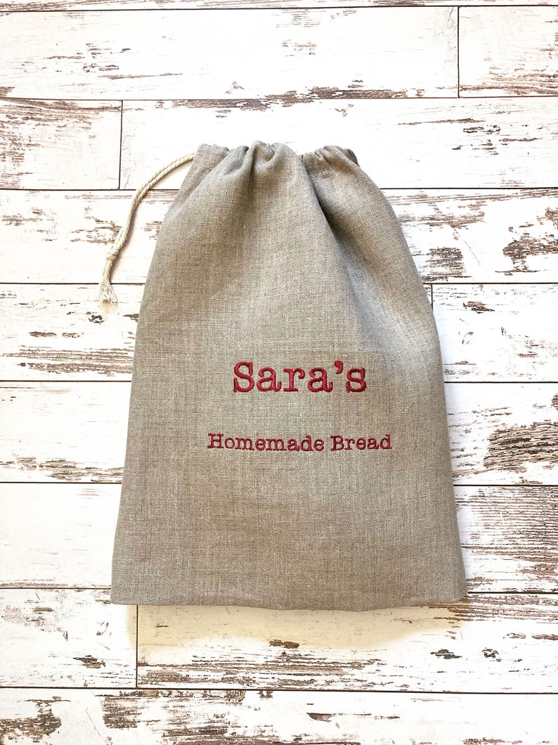 May include: A beige linen bread bag with a drawstring closure. The bag features red embroidered text that reads "Sara's Homemade Bread". The bag is designed to store homemade bread.