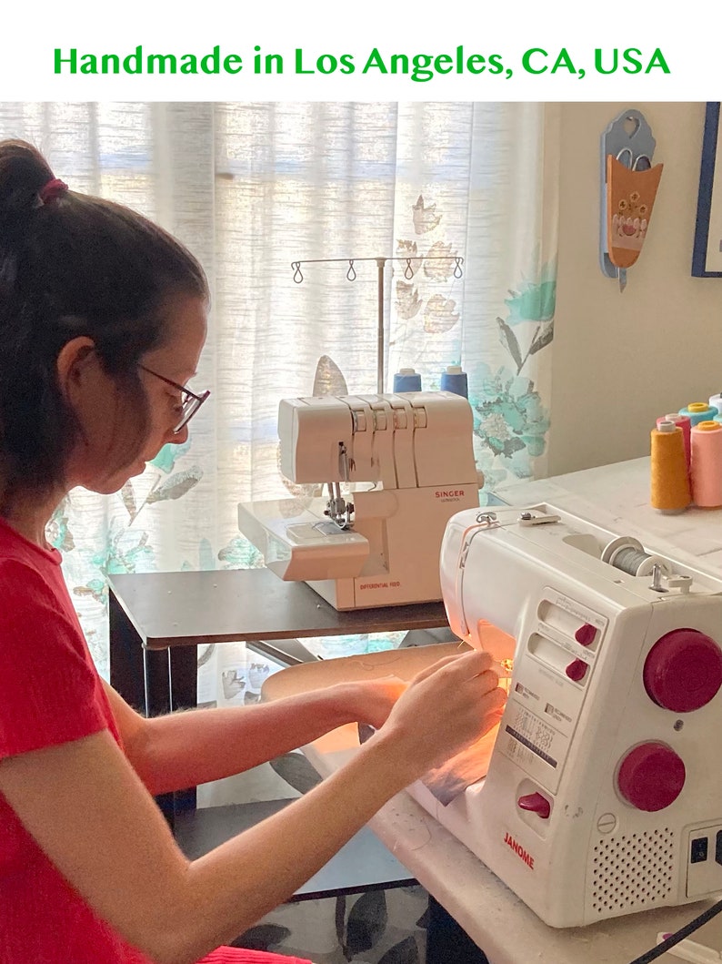 May include: A white Janome sewing machine with pink dials and a white Singer serger machine are positioned on a table. The text Handmade in Los Angeles, CA, USA is displayed at the top of the image. The sewing machines are used for fabric work.