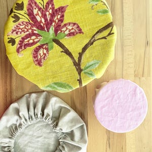 May include: Three round fabric bowl covers with elastic edges. One large cover features a yellow background with a large red and white floral print. One medium cover is solid light gray. One small cover is solid light pink.